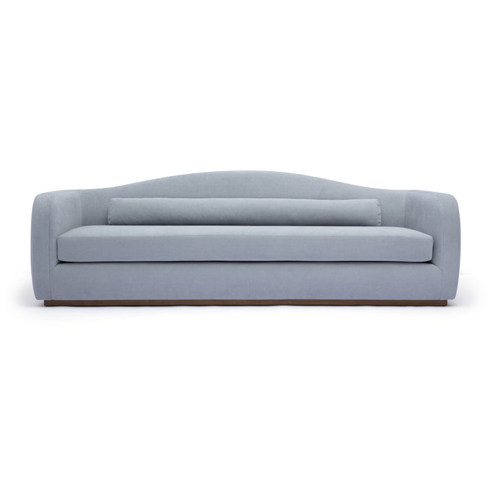 A modern three-seater sofa upholstered in a light blue-gray fabric, featuring a curved backrest, a matching lumbar pillow, and a low wooden base, shown against a plain white background.